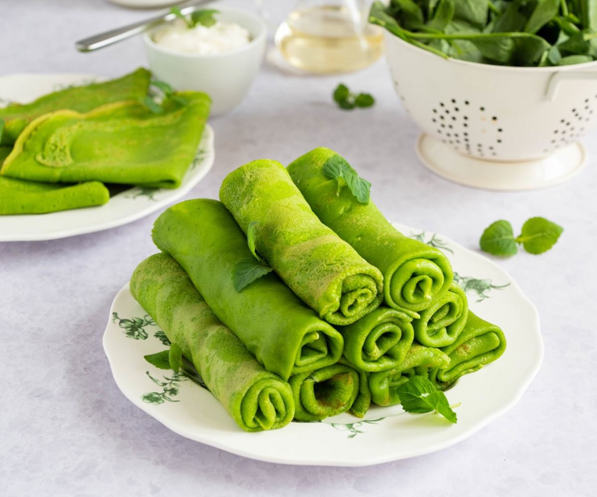 A plate of rolled spinach crepes with a vivid green color, garnished with fresh mint leaves and served with a dollop of ricotta in a bowl; additional crepes and a bowl of spinach in the background.
