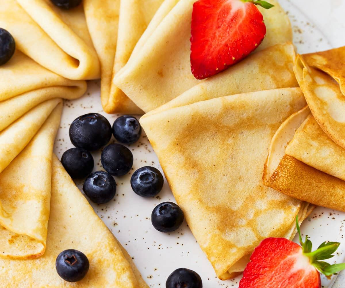 Freshly made golden crepes  following The Recipe Cloud recipe. Folded and served with fresh blueberries and sliced strawberries on a white plate.