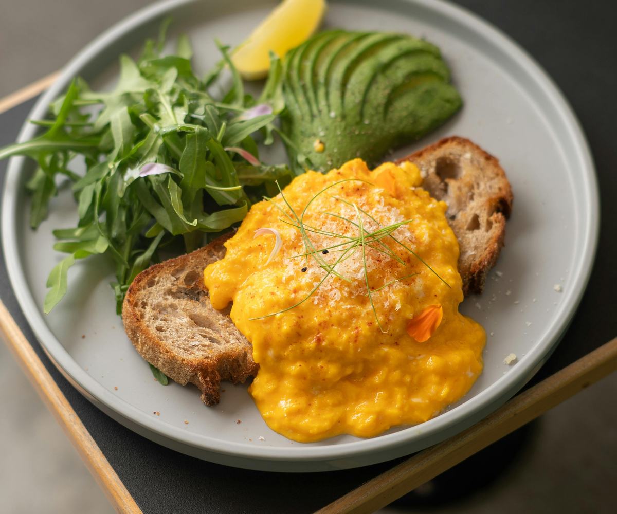Balanced plate with creamy scrambled eggs on crusty sourdough bread served alongside sliced avocado, fresh greens, and a lemon wedge.