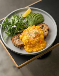 Balanced plate with creamy scrambled eggs on crusty sourdough bread served alongside sliced avocado, fresh greens, and a lemon wedge.