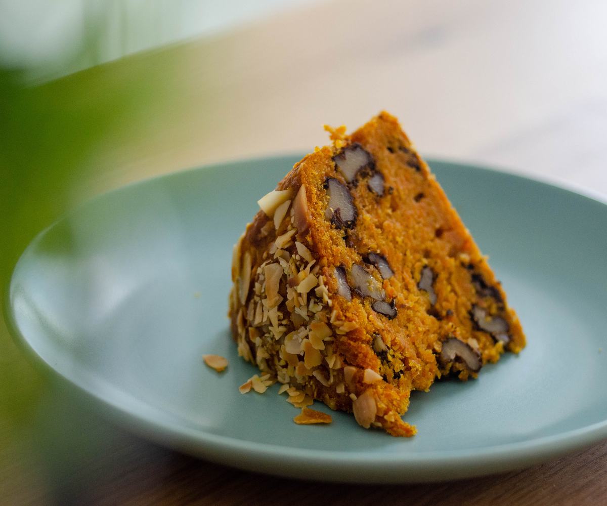 A close-up of a slice of moist carrot cake with walnuts and almond flakes on top, showing its rich orange color and tender texture, served on a light blue plate.