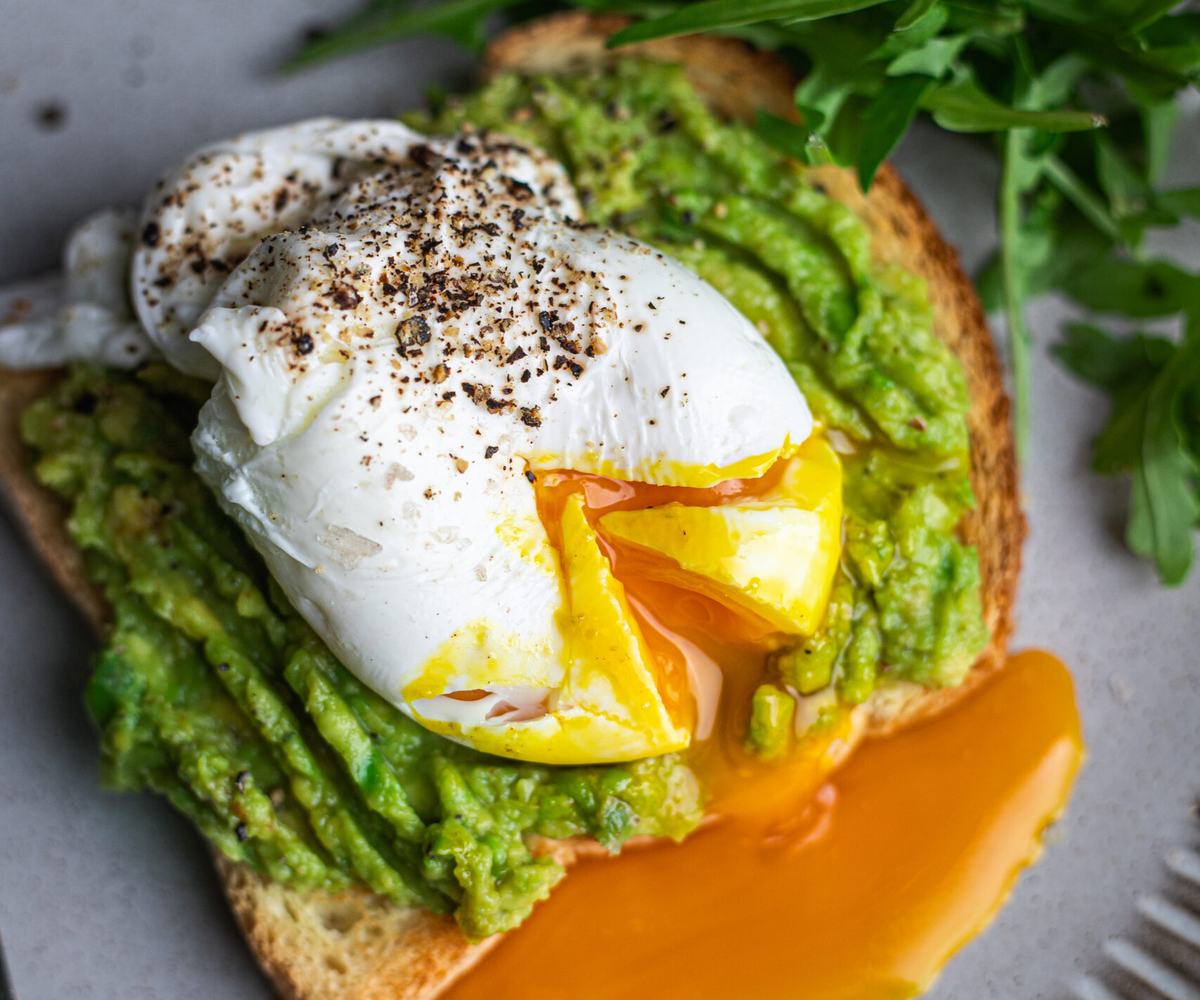 Freshly cooked for 3 minutes poached egg with runny yolk, served on a toasted slice of bread with mashed avocado, finished with fresh green herbs on a plate.