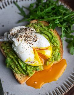 Freshly cooked for 3 minutes poached egg with runny yolk, served on a toasted slice of bread with mashed avocado, finished with fresh green herbs on a plate.