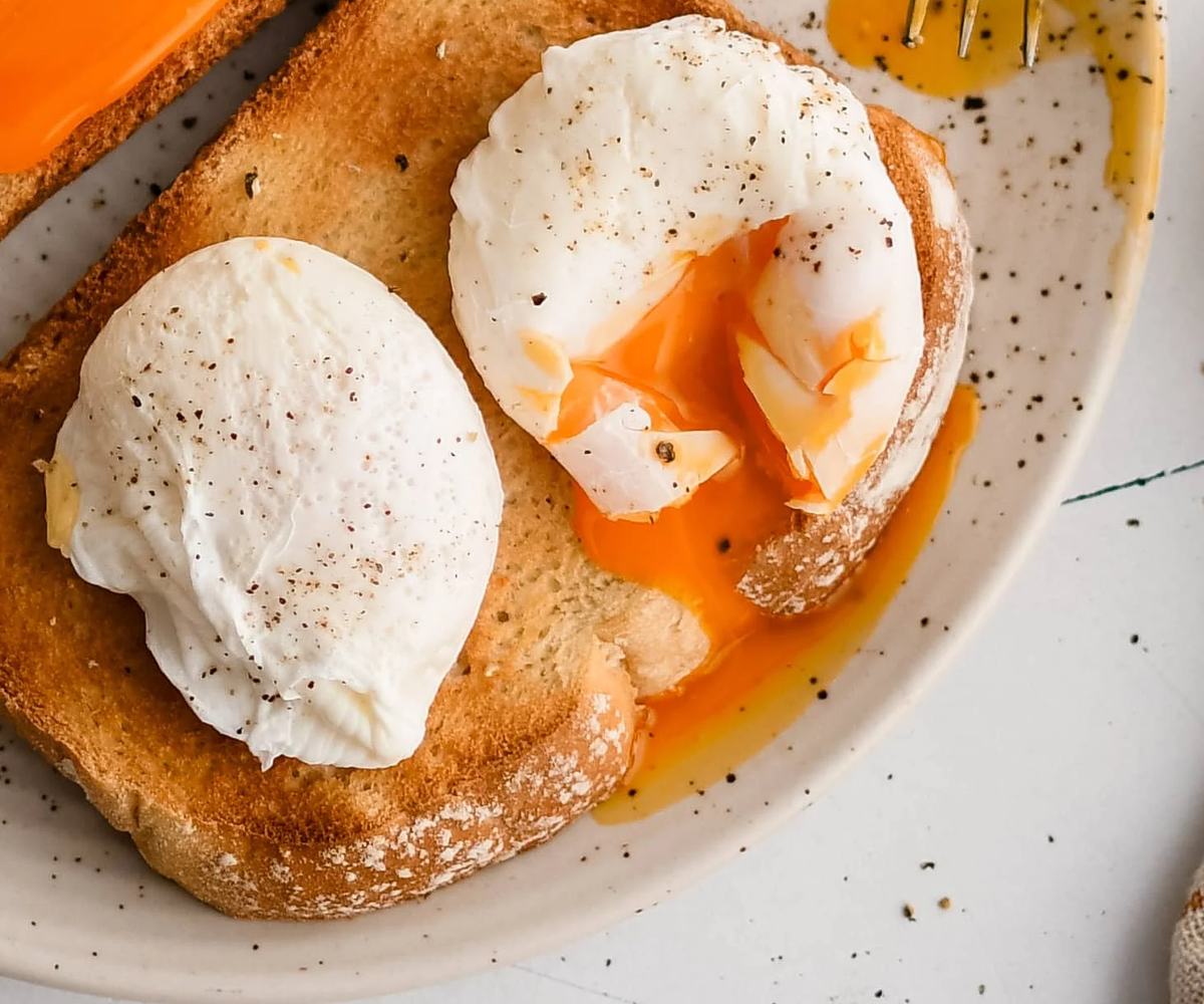 Two slices of toasted bread topped with poached eggs, cooked multiple at once, seasoned with black pepper, with runny yolks spilling onto the plate.